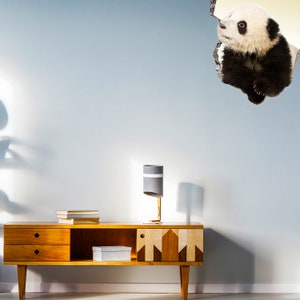 May include: A wooden media console with two drawers and an open shelf. A gray lamp with a white band sits on the console. A panda bear sticker is peeking out from a hole in the light blue wall behind the console.