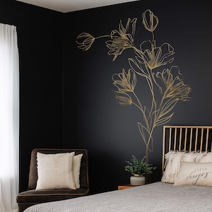 May include: A bedroom with a black wall adorned with a gold floral outline. A brown chair with white cushions is on the left, and a bed with a gold headboard is on the right. A small plant and decorative pillows complete the scene.