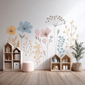 Puede incluir: Un adhesivo de pared que presenta una colección de flores silvestres dibujadas con líneas en tonos azul, rosa y amarillo. El arreglo floral incluye varios tallos, hojas y flores, exhibido sobre una pared lisa encima de estantes de madera.