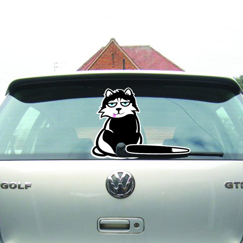 May include: A black and white cartoon cat sticker with a tongue sticking out, sitting on a car's rear window wiper blade. The cat has a white patch on its chest and a black tail.