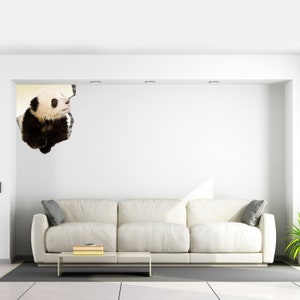 May include: A white sofa with two gray pillows sits in a white room with a white wall. A panda bear is peeking out from a hole in the wall.