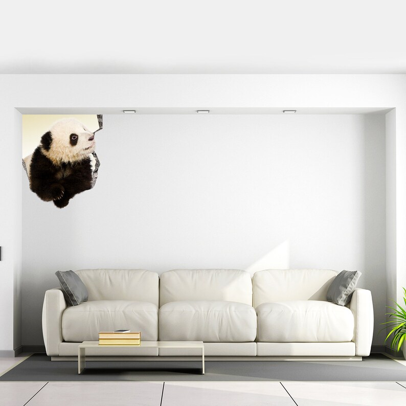 May include: A white sofa with two gray pillows sits in a white room with a white wall. A panda bear is peeking out from a hole in the wall.