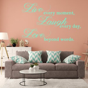 May include: A living room interior with a light brown couch and three pillows with a chevron pattern. The wall is a light peach color with a teal green vinyl decal that reads "Live every moment, Laugh every day, Love beyond words."