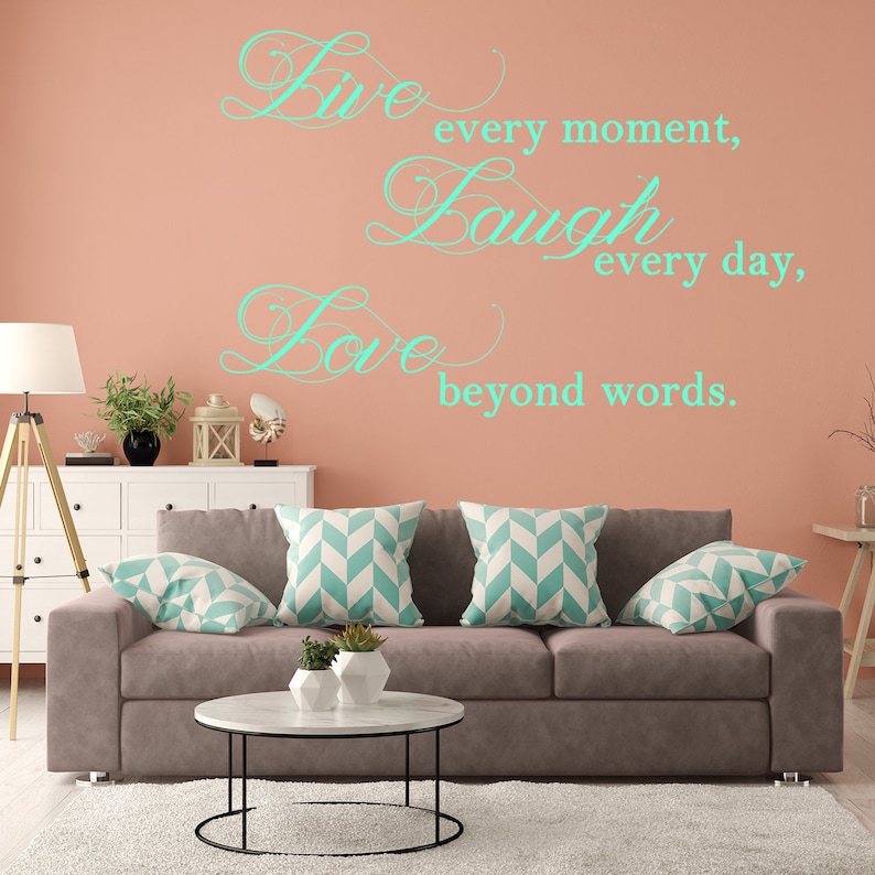 May include: A living room interior with a light brown couch and three pillows with a chevron pattern. The wall is a light peach color with a teal green vinyl decal that reads "Live every moment, Laugh every day, Love beyond words."