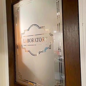 Könnte beinhalten: Eine Milchglastür mit einem dekorativen Rahmen und dem Wort "LABORATORY" in goldenen Lettern.