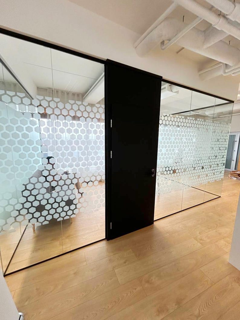 Puede incluir: Un espacio de oficina moderno con una partici&oacute;n de vidrio que presenta un patr&oacute;n de panal blanco. Una puerta negra es visible en el centro de la imagen. El suelo es de madera clara.