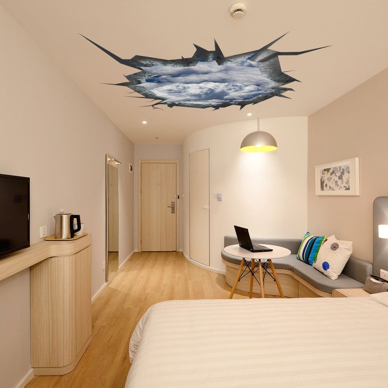 Puede incluir: Una calcoman&iacute;a de pared 3D que representa un techo agrietado con nubes visibles a trav&eacute;s del agujero. La calcoman&iacute;a se aplica a un techo blanco en una habitaci&oacute;n de hotel moderna.
