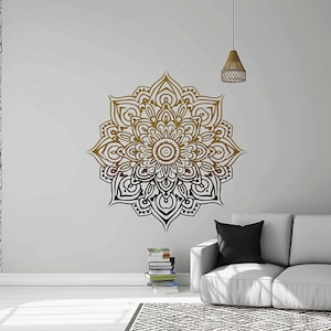 May include: A black and gold mandala wall decal with intricate details. The decal is applied to a white wall in a living room setting with a couch and a stack of books.