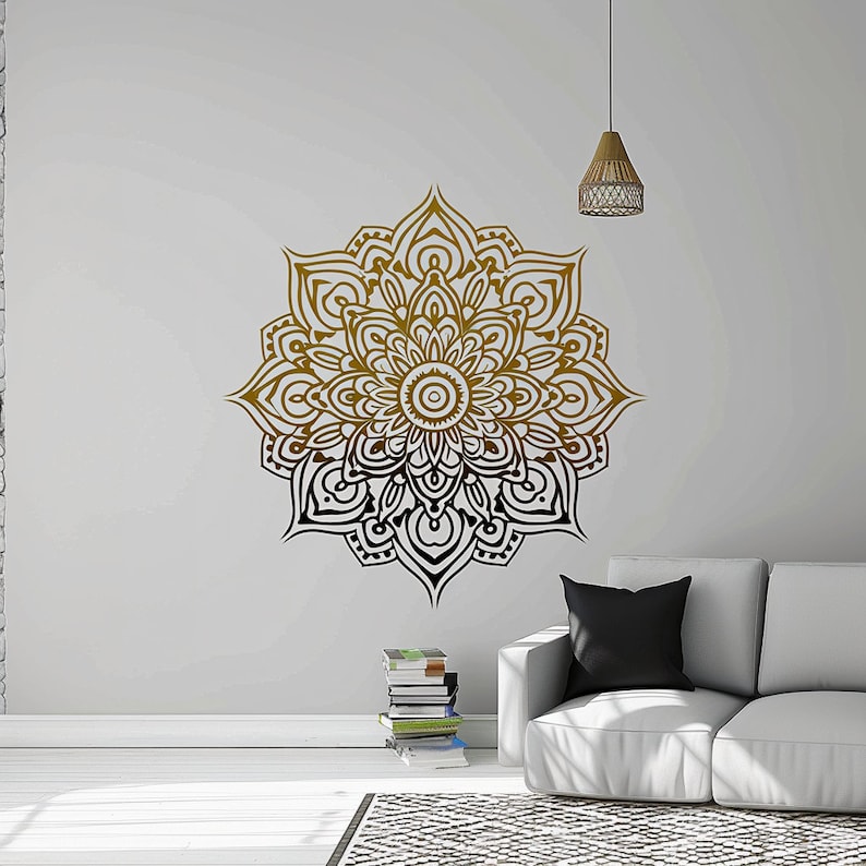 May include: A black and gold mandala wall decal with intricate details. The decal is applied to a white wall in a living room setting with a couch and a stack of books.