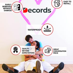 May include: A couple sits in front of a white wall with a pink heart painted on it. The heart has the word "Decords" written on it. There are several red circles with white icons and text inside them. The text includes "Easily Removed Without Damage", "Waterproof", "Easy to Install", "Quality Beyond Expectations", and "Great Conversation Piece".