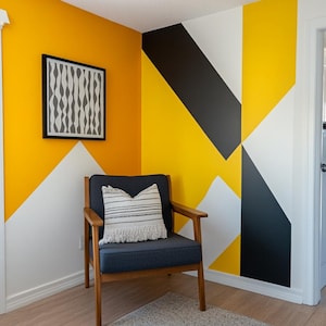 Op de afbeelding: Een woonkamer met een geel, zwart en wit geometrisch wandontwerp. Een grijze fauteuil met een wit en zwart gestreept kussen staat in de hoek van de kamer. Een grijs tapijt ligt op de vloer.