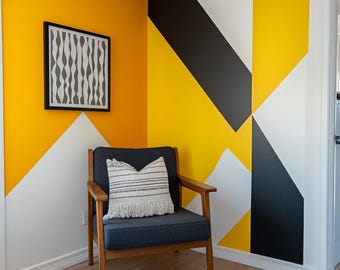 Puede incluir: Una sala de estar con un diseño de pared geométrico amarillo, negro y blanco. Un sillón gris con un cojín a rayas blanco y negro se encuentra en la esquina de la habitación. Una alfombra gris está en el suelo.