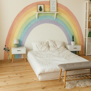 Op de afbeelding: Een wit bed met een wit dekbed en twee kussens staat voor een muur met een regenboog erop geschilderd. De regenboog is geschilderd in tinten oranje, geel, groen, blauw en paars. Boven het bed is een klein houten plankje met een ingelijst printje. Twee kleine houten nachtkastjes met lampen staan aan weerszijden van het bed. Een grijze bank staat voor het bed. De vloer is gemaakt van licht hout.