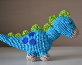 Op de afbeelding: Een blauw gehaakt dinosaurus speelgoed met groene stekels en blauwe vlekken. De dinosaurus heeft een lange staart en een klein hoofd met een bruine neus.
