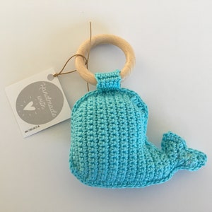 Op de afbeelding: Een gehaakt blauw walvis speelgoed bevestigd aan een houten bijtring. Het speelgoed heeft een label met de tekst "Handmade with love" en een website-adres.