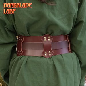 Pu&ograve; includere: Una cintura in pelle marrone con dettagli metallici decorativi e una fibbia. La cintura &egrave; indossata sopra una camicia verde.