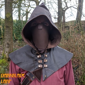 May include: A brown leather hooded cloak with a matching face mask. The hood has a drawstring closure and the cloak has a lace-up front with metal eyelets and buckles.