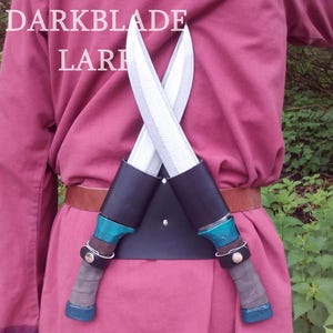 May include: Two silver LARP daggers with teal handles are crossed and secured to a black leather belt with a brown leather strap. The belt is worn over a pink tunic with the text "DARKBLADE LARP" printed on the front.