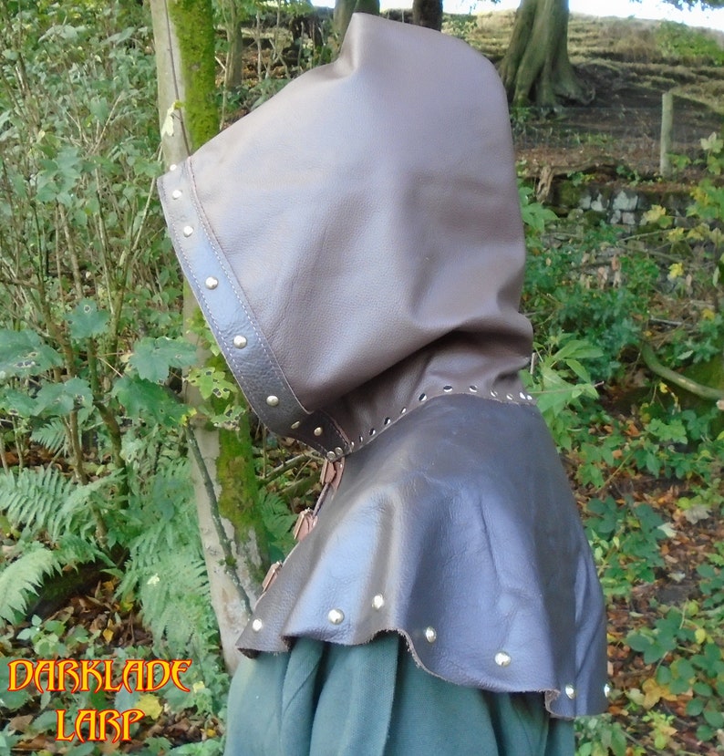 May include: A brown leather hooded cloak with a decorative trim and metal studs. The hood is pulled up and the cloak is worn over a green shirt.