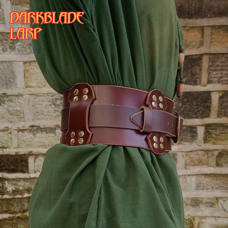 Pu&ograve; includere: Una cintura in pelle marrone con dettagli e fibbie in metallo dorato. La cintura &egrave; indossata sopra un capo di abbigliamento verde. Il testo "DARKBLADE LARP" &egrave; visibile nell'angolo in alto a sinistra dell'immagine.