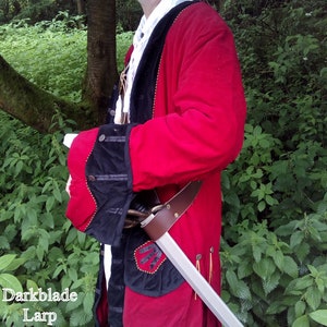 Pode incluir: Um casaco de pirata vermelho e preto com detalhes dourados e uma espada prateada presa ao lado. O casaco tem um colarinho e punhos de veludo preto.