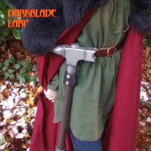 Op de afbeelding: Een persoon draagt een rode mantel en een groen shirt met een bruine leren riem en houdt een zilveren hamer vast met een donkerbruine houten steel. De hamer is met een leren riem aan de riem bevestigd.