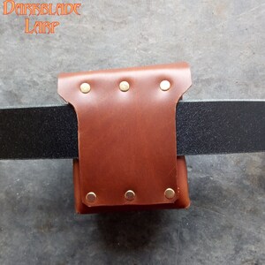 May include: A brown leather belt pouch with brass rivets. The pouch is attached to a black leather belt.