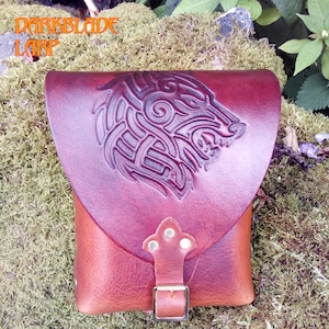 May include: A brown leather pouch featuring a flap with an embossed wolf design and a metal buckle closure. The text DARKBLADE LARP is visible in the upper left corner. The pouch has a smooth, reddish-brown finish on the flap and a textured body.