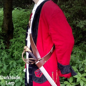 A Larp cutlass sheathed in a brown cross body abldric. The baldric is worn over a red and black frock coat and a white ruffled shirt.