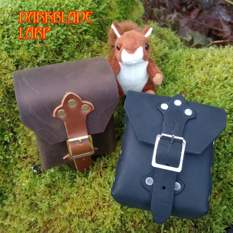 May include: Two leather pouches with buckles and straps. The pouch on the left is brown and the pouch on the right is black.  The pouches are sitting on a bed of green moss.  DARKBLADE LARP is written in orange text on a green background.