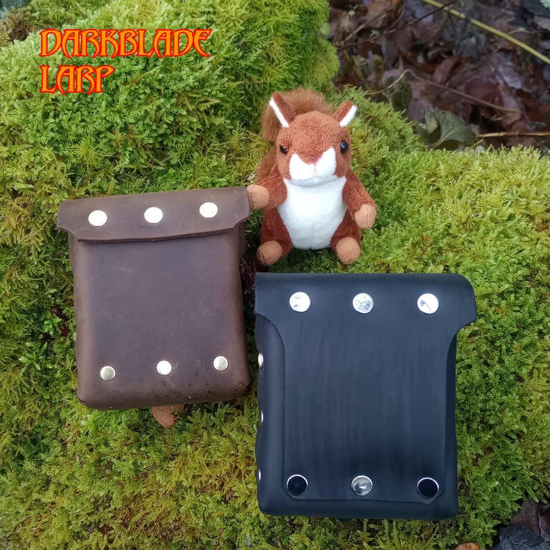 May include: Two leather pouches with metal rivets. The pouch on the left is brown and the pouch on the right is black.  DARKBLADE LARP is written in orange text on a green background.