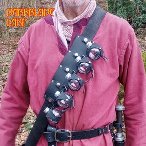 May include: A black leather shoulder harness with five glass vials, each held in a leather cup. The vials contain a red liquid. The harness is worn over a red tunic. The text "DARKBLADE LARP" is visible in the upper left corner.