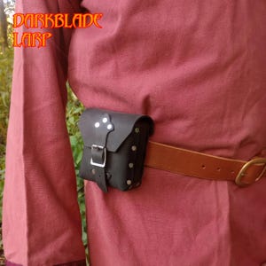 Könnte beinhalten: Ein schwarzer Lederbeutel mit einer silbernen Schnalle und Metallverzierungen. Der Beutel ist an einem braunen Ledergürtel befestigt.