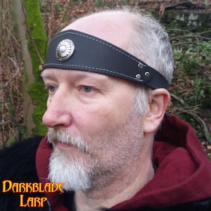 May include: A black leather headband with a silver metal circular design in the center. The headband has a stitched edge and silver metal rivets.