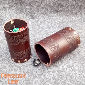 May include: Two brown leather dice cups with embossed Celtic knotwork and brass rivets. One cup contains colorful dice. The cups are cylindrical and have a rolled top edge. The Darkblade Larp logo is visible at the bottom.