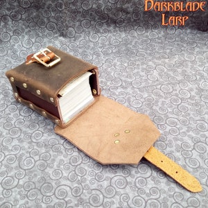 May include: A brown leather card holder with a flap and a buckle closure. The holder is open and a stack of cards is visible inside. The holder is on a gray and white patterned fabric.