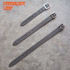 Pode incluir: Três tiras de couro preto com fivelas prateadas. As tiras são de comprimentos diferentes e têm vários furos para ajustar o ajuste.