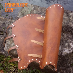 Op de afbeelding: Bruin leren LARP-pantser met de tekst "DARKBLADE LARP" in oranje. Het pantser heeft klinknagels en riemen om vast te maken. Het ontwerp lijkt te zijn voor een been of arm, met een gebogen vorm en een cilindrisch gedeelte.