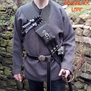 Op de afbeelding: Een zwart leren LARP (Live Action Role Playing) schouderharnas met een tasje, meshouders en een riem met een decoratieve gesp. De persoon draagt een grijze tuniek en houdt een zwaard vast.