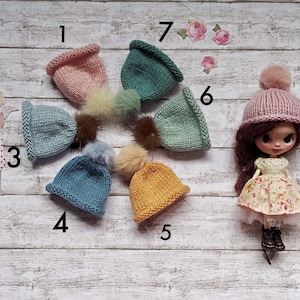 May include: Seven knitted hats with pom-poms in various colors, including pink, blue, green, and yellow. The hats are arranged in a circle on a white wooden background.