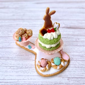May include: A tiered cake stand with a green cake topped with a chocolate bunny, a pink and white cake with a strawberry, and a plate of cookies. The stand is decorated with pastel pink and blue bunnies.