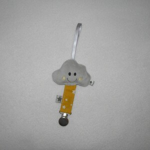 Peut inclure: Un jouet en peluche en forme de nuage gris avec un visage souriant et des yeux jaunes. Le jouet est attaché à un ruban jaune et blanc avec de petites étoiles blanches. Le ruban est attaché à un clip argenté.