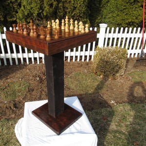 May include: A handcrafted wooden chess table with a dark brown finish. The square tabletop holds a complete chess set with light and dark pieces. The table is supported by a dark, square column and base, set outdoors near a white picket fence.