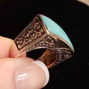 May include: A silver ring with a turquoise stone set in a square shape. The ring has an intricate design around the stone.