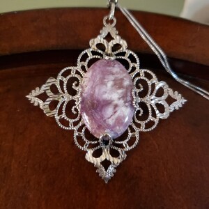 May include: A silver filigree pendant with a purple and white gemstone. The pendant is hanging from a silver chain.