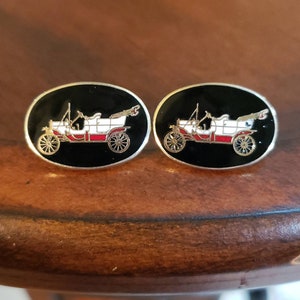 May include: A pair of cufflinks featuring a vintage car design. The car is white with red accents and is set against a black background. The cufflinks are oval shaped and have a silver finish.