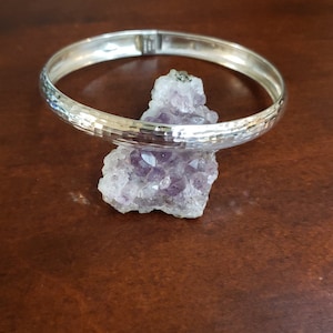 May include: A silver hammered bangle bracelet with a smooth, polished finish. The bracelet is displayed on a purple amethyst crystal.
