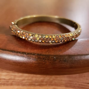 May include: A gold-toned bangle bracelet with a band of multi-colored rhinestones. The rhinestones are in shades of brown, orange, and yellow.