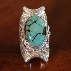 May include: A silver ring with an oval turquoise stone set in a detailed silver band. The band is decorated with intricate designs and a Greek key pattern.