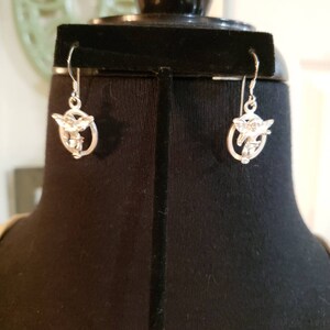 May include: A pair of silver dangle earrings featuring a cherub design. The cherubs are holding a heart and arrow.